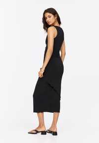 Schwarzes, figurbetontes Kleid mit Rundhalsausschnitt, gerafftem Oberteil und gewelltem Saum. Das Modell kombiniert es mit schwarzen, hochhackigen Sandalen.