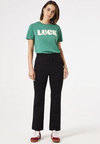 Groene katoenen t-shirt met vetgedrukte witte tekst "LUCK", gecombineerd met zwarte hoge taille broek en rode sneakers met witte veters.