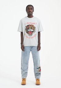 Světlé šedé oversized tričko se živou grafikou tygra a textem "Ed Hardy Los Angeles". Nosené s vybledlými modrými džíny a béžovými botami.