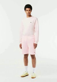 Mladý muž stojí oblečený v svetloružovej mikine a zodpovedajúcich šortkách s bielymi ponožkami a béžovými teniskami na bielom pozadí.