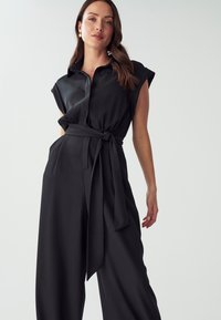 Zwart jumpsuit met korte mouwen, een knoopsluiting aan de voorkant en een tailleband. Gemaakt van een soepele stof met wijde broekspijpen.
