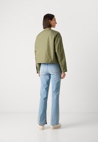 Giacca bomber corta verde oliva, abbinata a jeans a zampa d'elefante blu chiaro e sneakers bianche. Tessuto morbido con dettagli di bottoni e tasche.