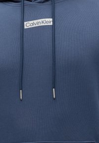 Μπλε φούτερ με κουκούλα με το λογότυπο Calvin Klein σε λευκό στο στήθος και ταιριαστά μπλε κορδόνια με ασημένιες μεταλλικές άκρες.