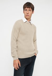 Beige geribbeld sweater met een ronde hals, textuur brei ontwerp en lange mouwen, gecombineerd met donkere denim jeans. Witte blouse is zichtbaar.