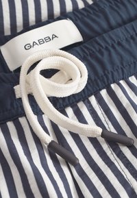 Tkanina mornarskih prugica s bijelim užetom za vezanje, crnim plastičnim završetcima i bijelom etiketom na kojoj piše "GABBA." Prikaz užeg dijela pojasa.