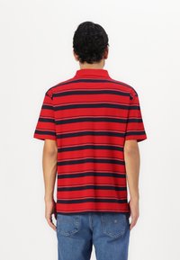 Polo rouge avec des rayures horizontales bleu marine, manches courtes et col classique. Le tissu semble lisse avec une texture légère.