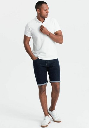 Weißes Poloshirt, dunkelblaue Jeansshorts mit aufgerollten Säumen und weiße Sneakers. Das Outfit zeichnet sich durch ein lässiges, tailliertes Design aus.