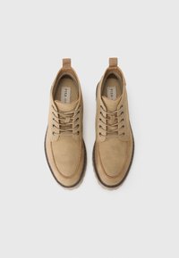 Zapatos de ante beige con una textura suave, punta redonda, costuras en contraste y cierre de cordones. Suela de goma con un lazo en la parte posterior.