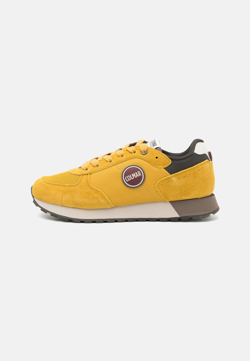 Sneakers din suede galben cu un logo rotund, accente maro, guler captusit și talpă din cauciuc. Șireturile și detaliile de cusătură sunt proeminente.