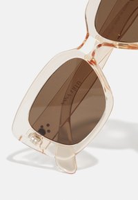 Transparente pfirsichfarbene Sonnenbrille mit großen braunen Linsen. Besitzt eine markante geometrische Form und subtile Hardware-Details.