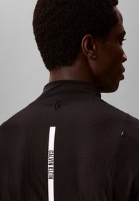 Zwarte persoon draagt een gestructureerde zwarte top met een wit verticaal Calvin Klein-logo op de rug, zijwaarts kijkend tegen een effen achtergrond.