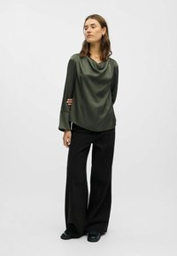 Oliven grøn cowl-neck top med lange ærmer, draperet stof, parret med højtaljede sorte breddebukser og sort fodtøj.