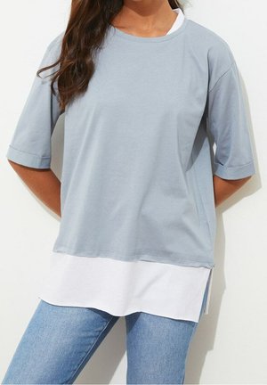 Kobieta ubrana w jasnoniebieską luźną bluzkę z krótkim rękawem nałożoną na dłuższą białą koszulę, w połączeniu z jasnoniebieskimi dżinsami, stoi na białym tle.