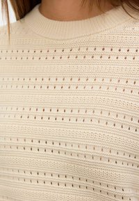 Pull en maille beige avec des rangées horizontales de petits trous en œillets et des motifs texturés, porté par une personne.
