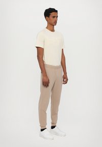 Crème t-shirt met reliëflogo, gecombineerd met beige sweatpants. Model draagt witte sneakers; algehele casual sportieve stijl en pasvorm.