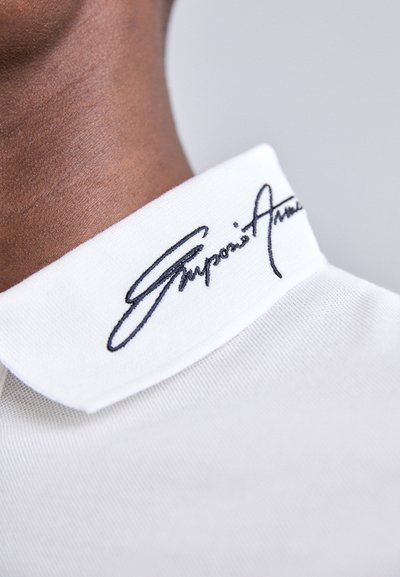 Hvit polo-skjorte med krage som har brodert svart signatur. Stoffet er teksturert, med en glatt overflate og rene kanter.