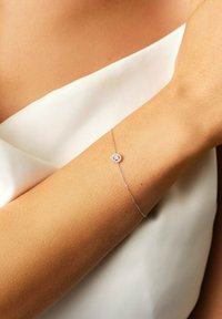 Delicate silver chain bracelet with a small circular diamond centerpiece worn on a woman’s arm against a white fabric background.
