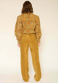 Femme aux cheveux bruns mi-longs portant un chemisier à motifs cachemire moutarde et un pantalon en velours côtelé moutarde taille haute et jambes larges, debout de dos.