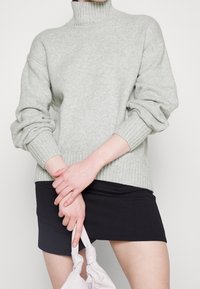 Jasnoszary sweter z golfem z prążkowanymi mankietami i dolnym brzegiem, w połączeniu z czarną mini spódniczką. Modelka trzyma jasną torebkę.