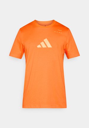 Πορτοκαλί αθλητική μπλούζα με κοντά μανίκια, με μεγάλο λογότυπο με τρεις λωρίδες ανοιχτό πορτοκαλί στο στήθος και λεπτομέρεια κειμένου στον ώμο.