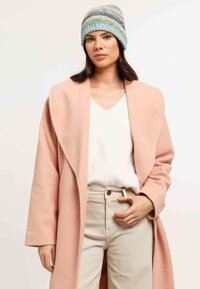 Donna che indossa un cappotto rosa pastello, una maglietta bianca, pantaloni beige e un berretto lavorato a maglia blu-verde, in posa su uno sfondo neutro.