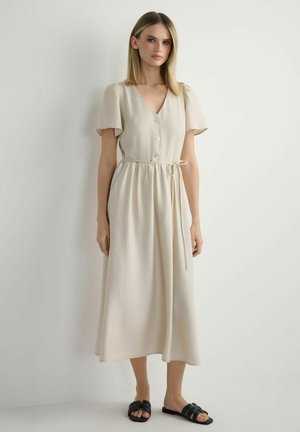 Kobieta stoi przy białej ścianie, ubrana w beżową sukienkę midi z guzikami, krótkimi rękawami, wiązaniem w talii oraz czarne, otwarte, płaskie sandały.