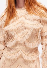Beige mekko, jossa on kerrokselliset röyhelöt ja kukkakirjonta. Sisältää läpikuultavaa kangasta, jossa on kimalteita ja teksturoitu pinta. Elegantti ja monimutkainen muotoilu.