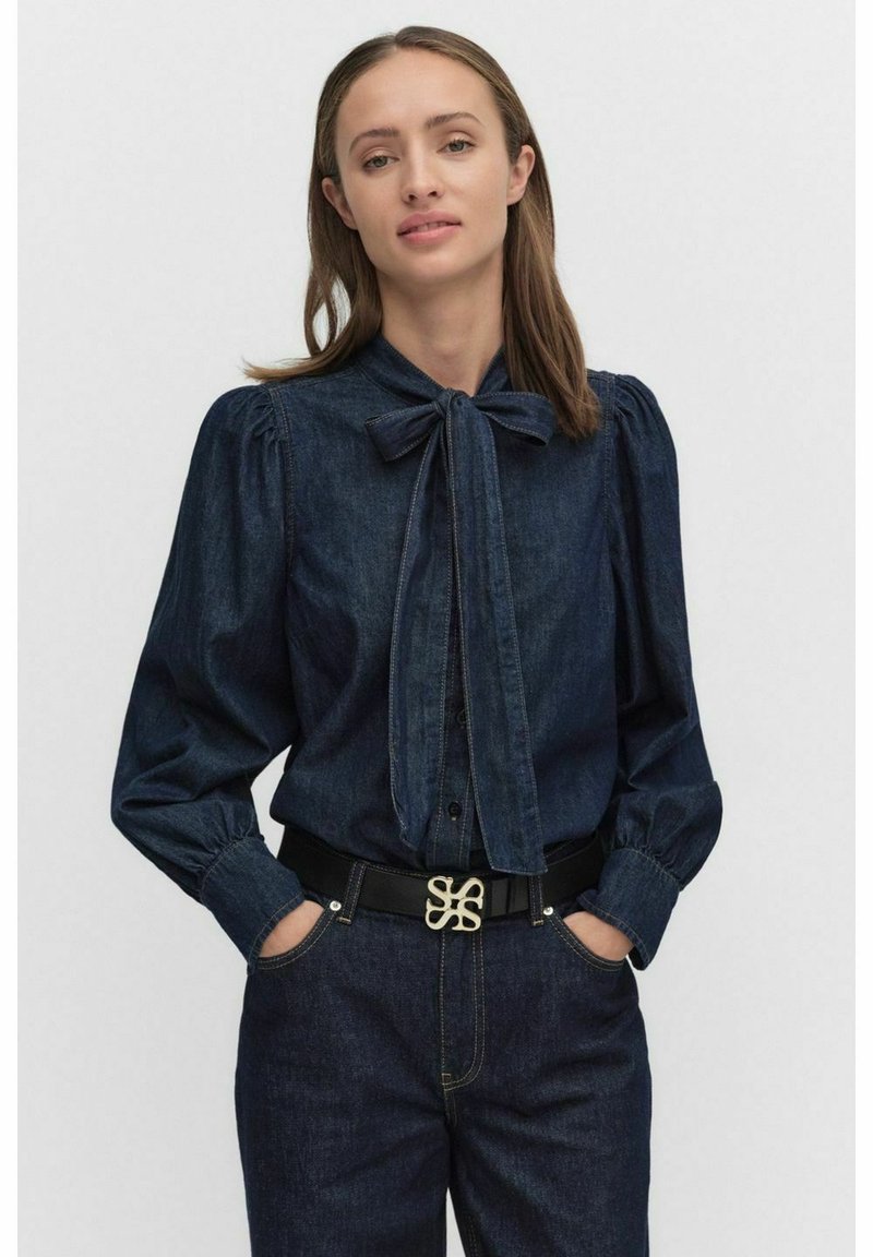 Mörk denimskjorta med en knuten rosettkrage, puffärm och knappar framtill. Matchad med högmidjade denimbyxor och ett svart bälte med en dekorativ spänne.