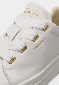 Λευκό δερμάτινο αθλητικό παπούτσι με χοντρές λευκές κορδόνες, χρυσά μεταλλικά τρύπια, και το λογότυπο "GANT" σε χρυσό στο πλάι και στη γλώσσα.