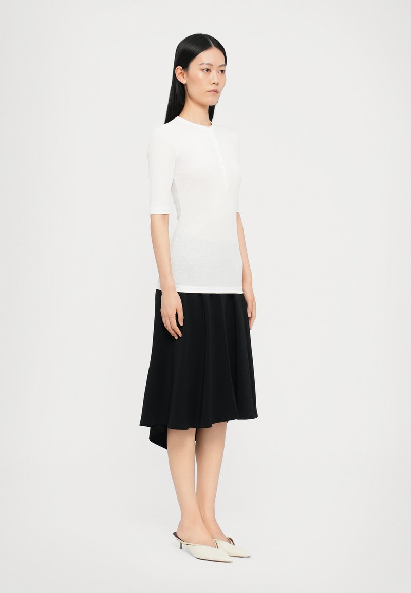 Top bianco aderente con maniche corte e scollatura tonda, abbinato a una gonna nera a ruota e mule di colore chiaro. Texture lisce visibili.