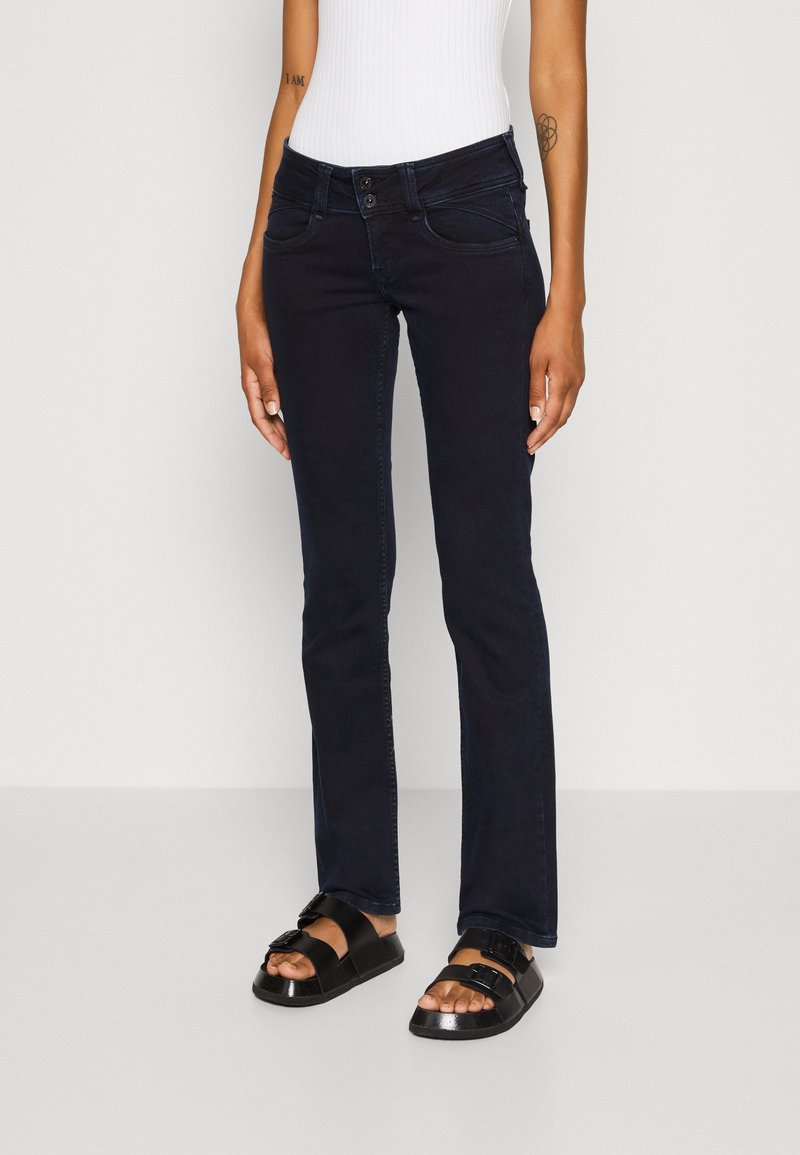 Tmavě modré zvonové džíny s pěti kapsami, zapínáním na knoflík a zip a jemnými švy; v kombinaci s černými sandály.