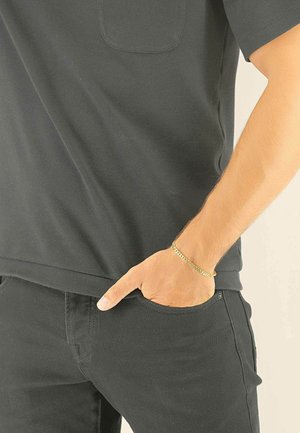 Chemise noire à manches courtes avec une poche poitrine, portée avec un pantalon noir. Un bracelet chaîne en or est porté au poignet, la main reposant dans la poche.