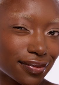 Gros plan d'un visage avec une peau brun foncé, texture lisse, lèvres brillantes et maquillage subtil. Les sourcils sont décolorés et de couleur claire.