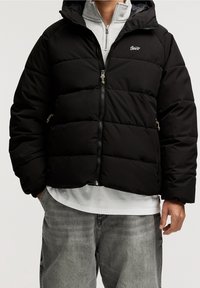 Chaqueta acolchada negra con capucha, cremallera frontal y bolsillos laterales. Presenta un logo en el pecho y una textura lisa y brillante.