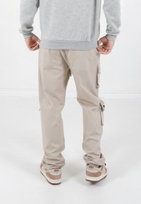 Beige Cargo-Hosen mit mehreren Taschen, Reißverschlussdetails und einem lockeren Schnitt, kombiniert mit tanfarbigen und braunen Nike-Sneakern. Grauer Pullover ist sichtbar.