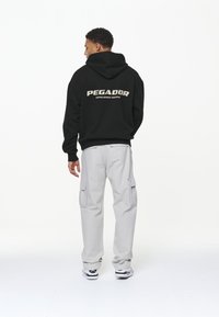 Sweatshirt negru cu glugă și logo-ul "PEGADOR" pe spate, asortat cu pantaloni cargo gri deschis, dotati cu buzunare laterale și adidași.