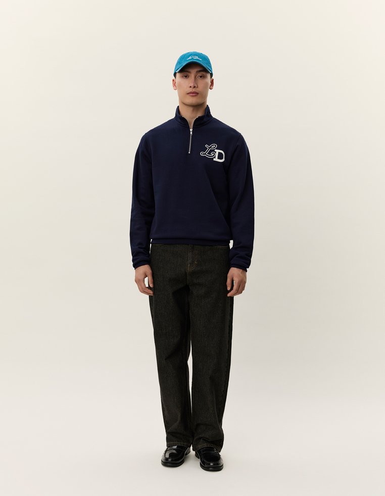 Felpa con zip blu marina con logo bianco, abbinata a pantaloni in denim scuro e un cappellino turchese. Il modello è in piedi contro uno sfondo neutro.