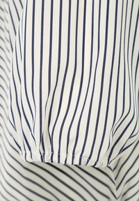 Tissu blanc avec des rayures verticales bleu marine, présentant un détail de manche froncée. Le matériau semble lisse et léger.