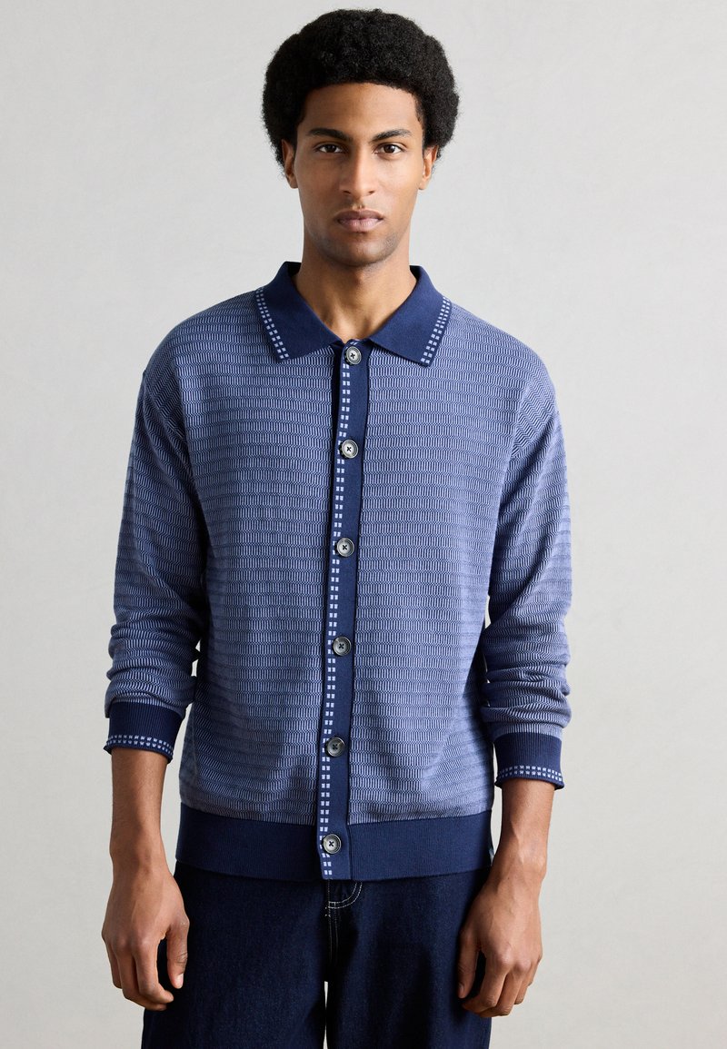 Ζακέτα με κουμπιά, πλεκτή σε υφή σε σκούρο μπλε χρώμα. Διαθέτει αντίθετο γιακά και μανσέτες σε ναυτικό μπλε με ανοιχτό μπλε λεπτομέρειες και κουμπιά.