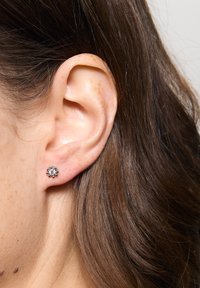 Srebrne kolczyki w kształcie sztyftów z okrągłym, przezroczystym kamieniem umieszczonym w kwiecistej oprawie, na tle teksturowanego ucha.