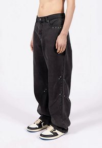 Osoba w luźnych czarnych spodniach z metalowymi oczkami i czarno-białych sneakersach, stojąca bokiem na białym tle.