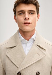 Beiger Fischgrät-Peacoat mit breiten Revers, doppelreihigem Knopfverschluss und einer strukturierten Stoffoberfläche über einem weißen Hemd mit Kragen.