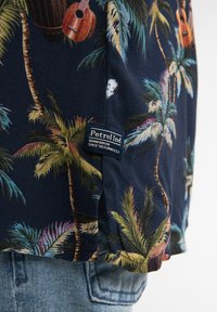 Cămașă scurtă de culoare bleumarin cu imprimeu tropical ce include palmieri, chitare și accente colorate. Include un eticheta "Petrol Ind" la margine.