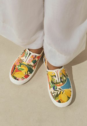 Nohy oblečené v bílých kalhotách a bezkrokových plátěných botách s barevným tropickým ovocným a listovým potiskem na béžové podlaze.