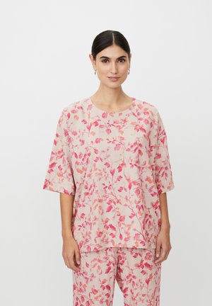 Jasnoróżowa bluzka w kwiatowy wzór w odcieniach czerwieni i koralu. Luźny krój, okrągły dekolt i rękawy do łokcia. Widoczne dopasowane spodnie.