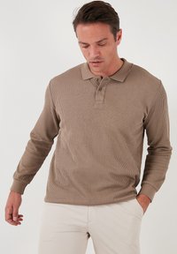 Polo a maniche lunghe in tessuto a coste marrone chiaro, con colletto, pattina con tre bottoni e polsini aderenti. Abbinato a pantaloni di colore chiaro.
