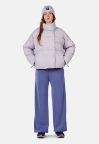 Helllila Steppjacke mit hohem Kragen, gestepptem Design und Fronttaschen. Kombiniert mit locker sitzenden blauen Sweatpants und einer passenden Mütze.
