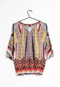 Blouse ample à motifs multiples avec des couleurs vives jaune, rouge et noir ; présente des motifs géométriques et floraux audacieux, des manches larges et un ourlet froncé.