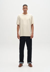 Beige Baumwoll-T-Shirt mit rundem Halsausschnitt und oversized Passform. Getragen mit dunkelblauen Jeans und braunen Lederschuhen im Bootsstil.