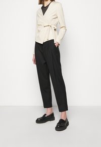 Beige obalový top se zavazováním v pase, spárovaný s černými plisovanými kalhotami a černými mokasíny. Top má design s výstřihem do V.